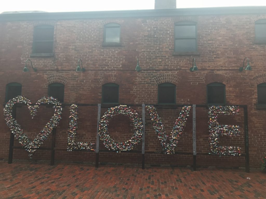 A collection of padlocks that spell out the word LOVE and form a heart against the backdrop of a brick building.