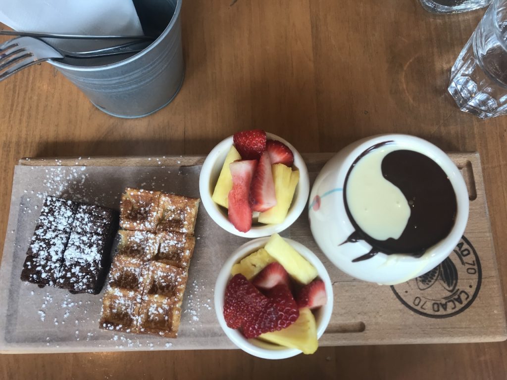 A powdered-sugar dusted brownie and waffle, two bowls of strawberries and pineapple, and a white bowl containing white chocolate and milk chocolate in the shape of a ying yang sign, all on a wooden tray shot from above.