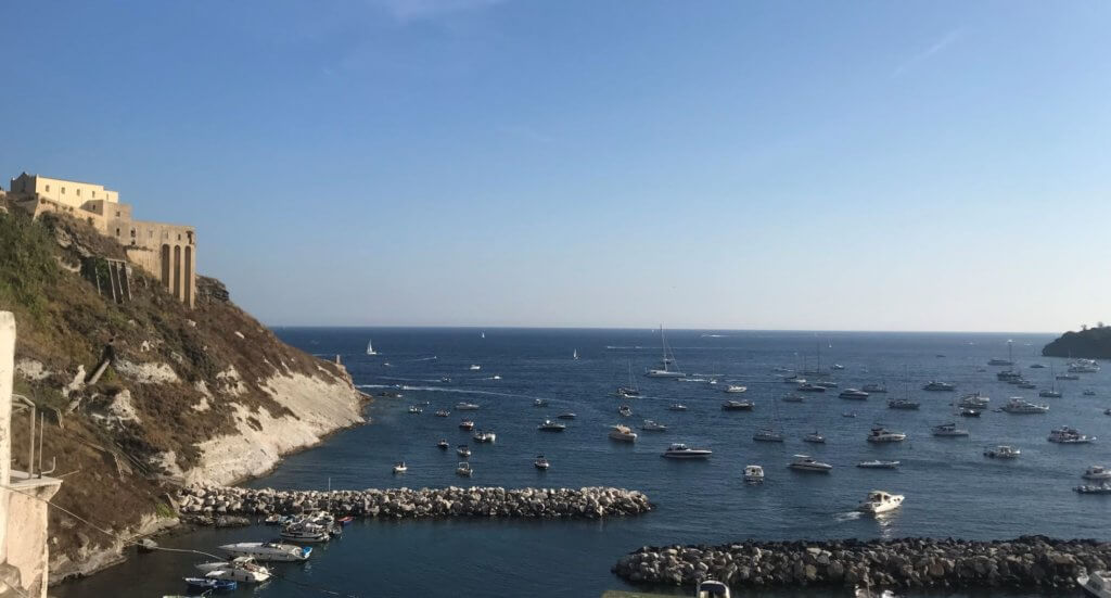 Distant boats dot the dark blue water, and buildings are visible on the side from an overlook point.