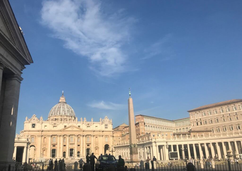Pale, intricately designed buildings framing a plaza with a blue sky overhead. Occasional people are spread out across the plaza.