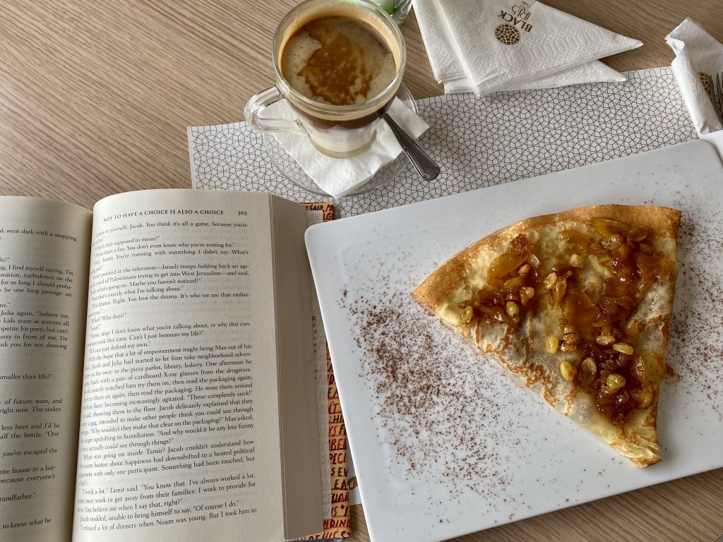 A half-open book on a table next to an apple crepe sprinkled with nuts and cinnamon and a nearly-full cup of a hot chocolate coffee blend. This is one of the books the author read in 2020.