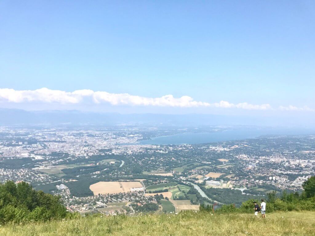Pale blue, cloudy skies stretch across the top part of the frame. Below, there are distant buildings, trees, and a lake. From this viewpoint, you can see the international city of Geneva from far out.