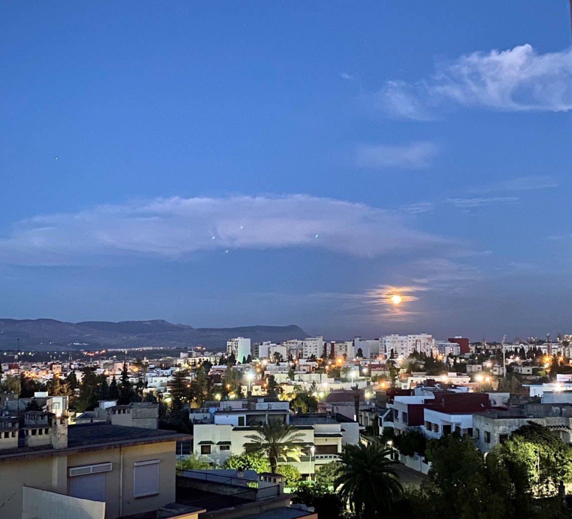 Lit-up buildings fill the bottom of the frame, while above, a pure blue sky with an almost-full moon marks the turn from day to night.