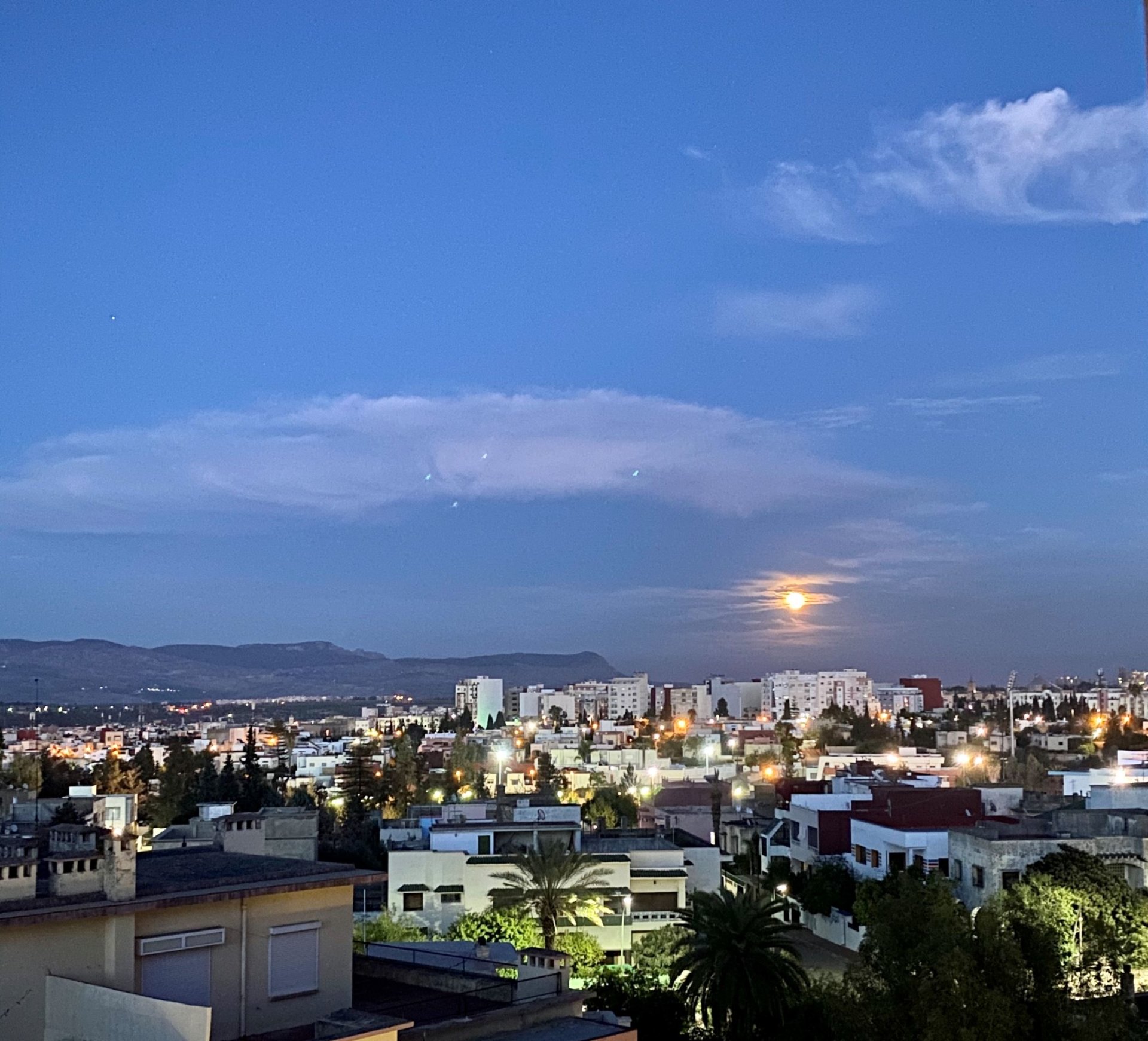 Lit-up buildings fill the bottom of the frame, while above, a pure blue sky with an almost-full moon marks the turn from day to night.