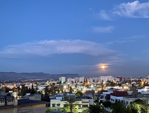 Lit-up buildings fill the bottom of the frame, while above, a pure blue sky with an almost-full moon marks the turn from day to night.