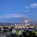 Lit-up buildings fill the bottom of the frame, while above, a pure blue sky with an almost-full moon marks the turn from day to night.